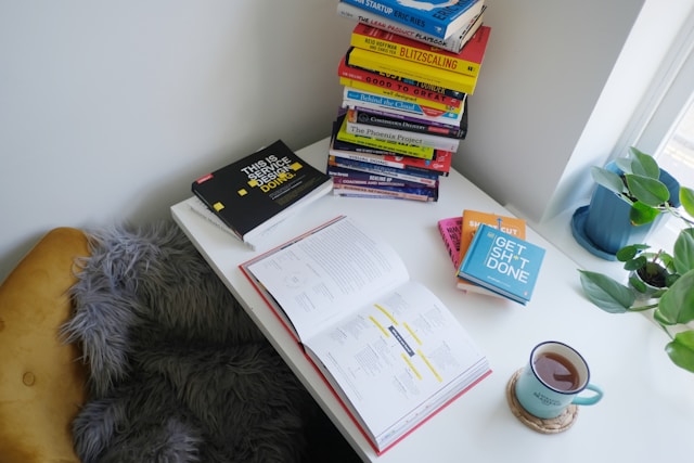 Desk by a window with business and software books in a stack with a mug of tea to the side.